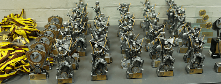 A collection of silver and gold cricket-themed trophies arranged in rows on a table. The trophies feature a cricket player in a batting stance with a shield-like background. The base of each trophy has a gold nameplate with engraved text.