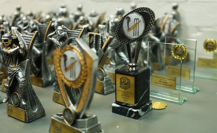 A close-up of various cricket trophies, including individual player awards and a larger championship trophy with a circular emblem featuring cricket equipment. The trophies are metallic silver with gold accents and have engraved nameplates. Transparent glass awards are also visible in the background.